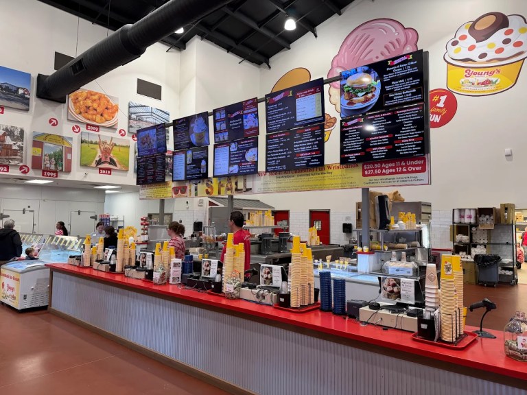 Interior view of a food stall featuring a brightly colored counter with ice cream cones and menu boards displaying various food items. Two employees are serving customers, and the walls are decorated with large images of food and desserts.
