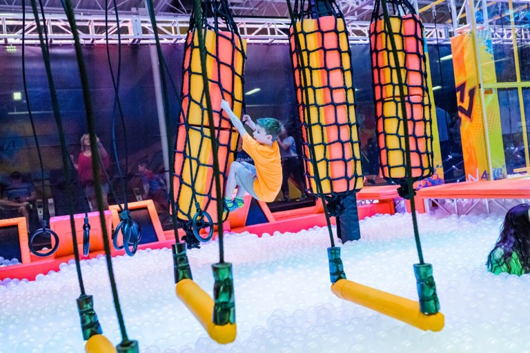 A child wearing an orange shirt climbs on a colorful obstacle in a play area filled with soft foam balls.