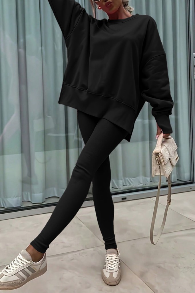 A person posing in a relaxed outfit consisting of a black oversized sweatshirt and black leggings, paired with white sneakers. They are standing in front of a sheer curtain backdrop.