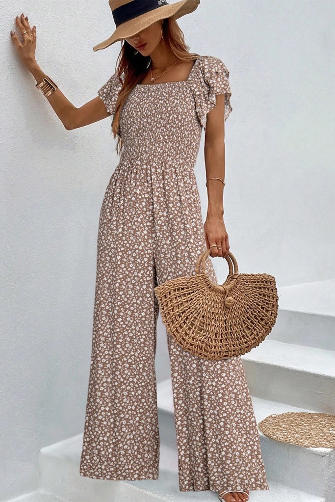 A woman wearing a floral print brown jumpsuit with ruffled sleeves, holding a woven straw handbag while standing on white stairs.
