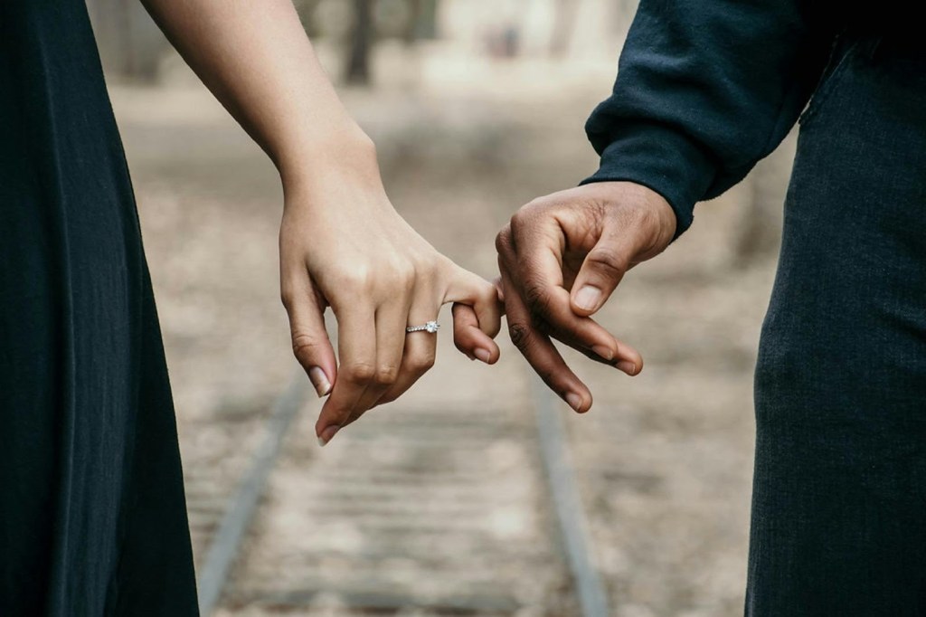 Close-up of two hands holding each other, one adorned with a sparkling ring, set against a blurred outdoor background.