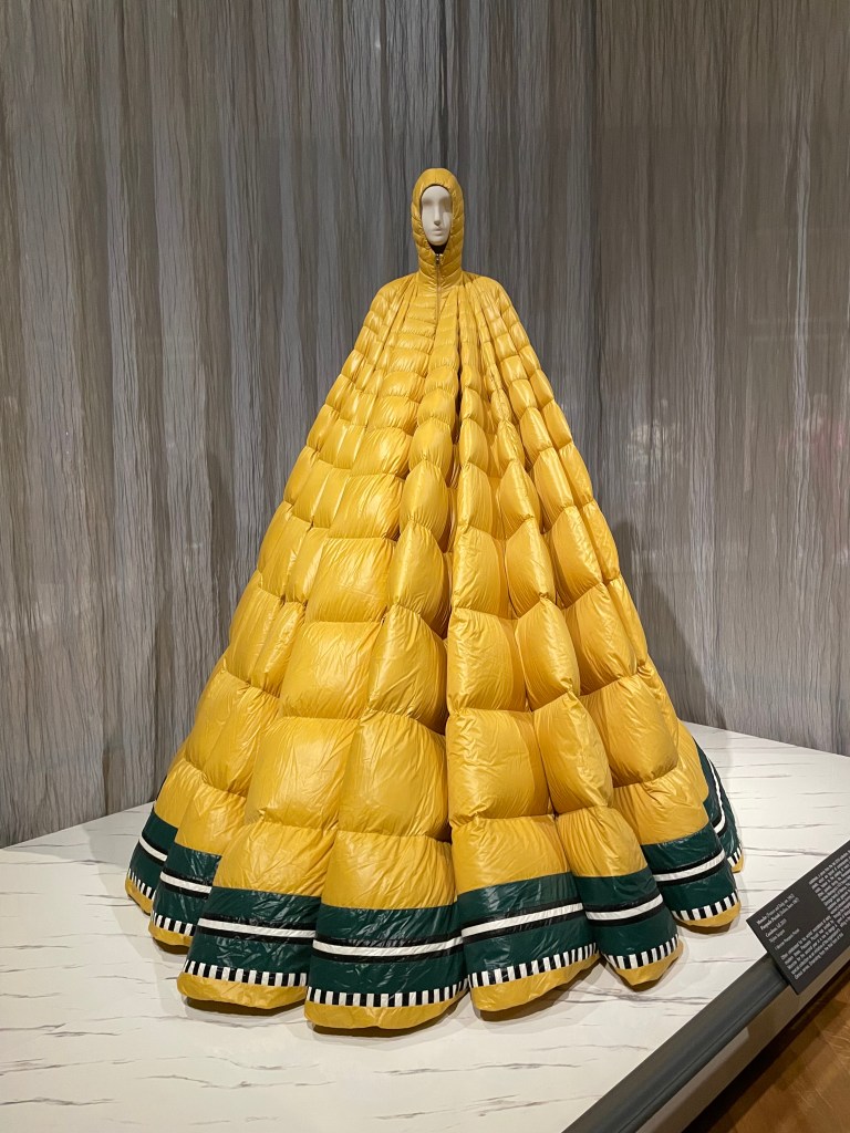 A unique, oversized yellow puffy garment displayed on a mannequin, featuring a hood and a voluminous shape with green and black striped accents at the hem.