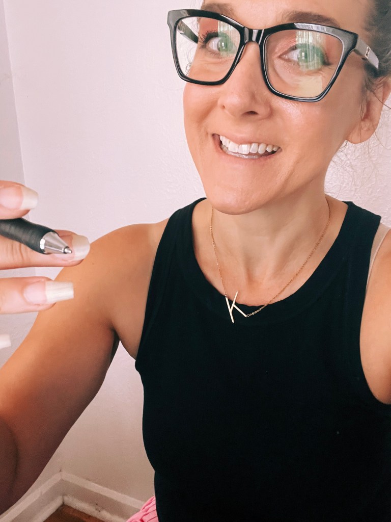 A woman with glasses smiling and holding a pen, wearing a black tank top and a gold necklace, sitting indoors.