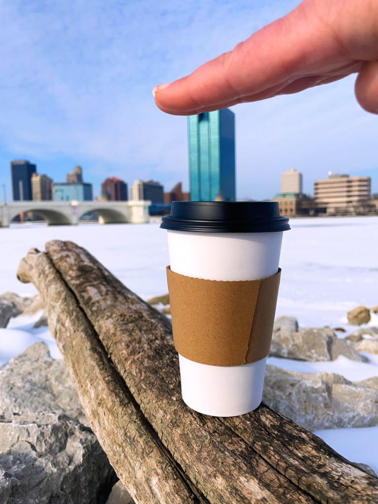 A hand playfully reaching towards a coffee cup sitting on a log, with a city skyline in the background and snow on the ground.