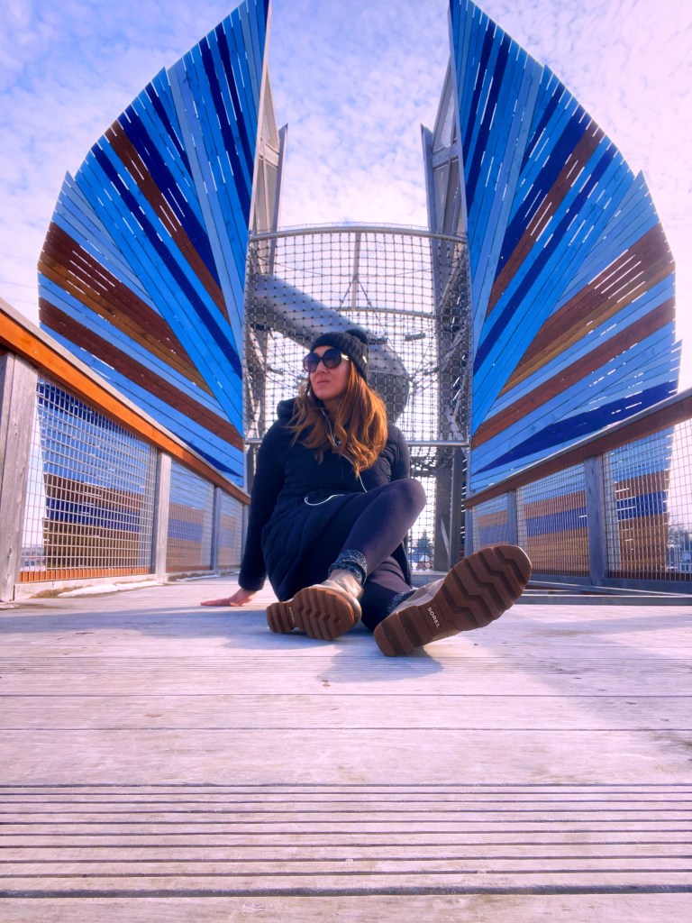 A woman sitting on a wooden walkway near a modern structure with blue and brown wooden panels resembling sails, surrounded by a scenic sky.
