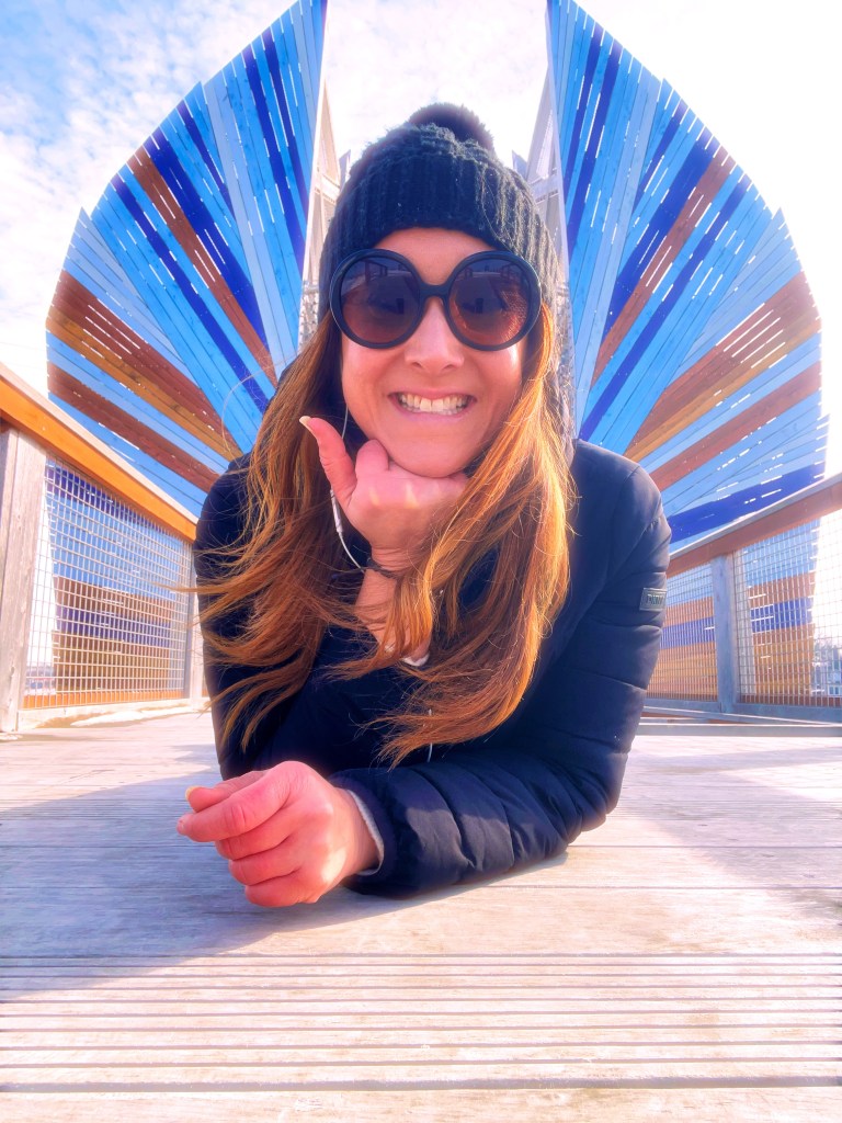 A woman with long hair, wearing sunglasses and a black beanie, smiles while resting her chin on her hand on a wooden surface. Behind her is a colorful, artistic structure made of blue and brown wooden slats.