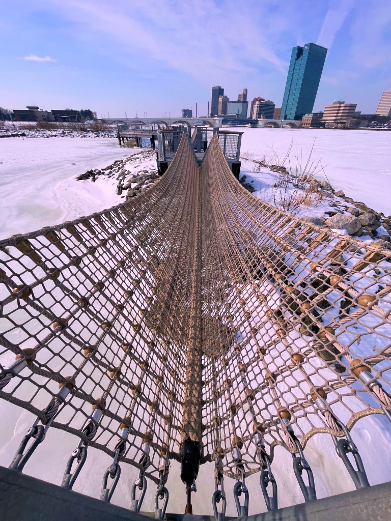A suspension bridge made of netting stretches over a frozen river, with a city skyline visible in the background under a clear blue sky.