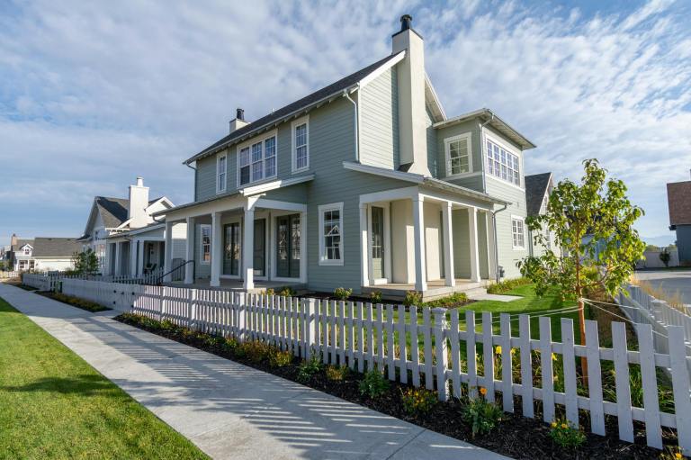 A modern green house with white trim and a front porch, surrounded by a manicured lawn and a white picket fence.