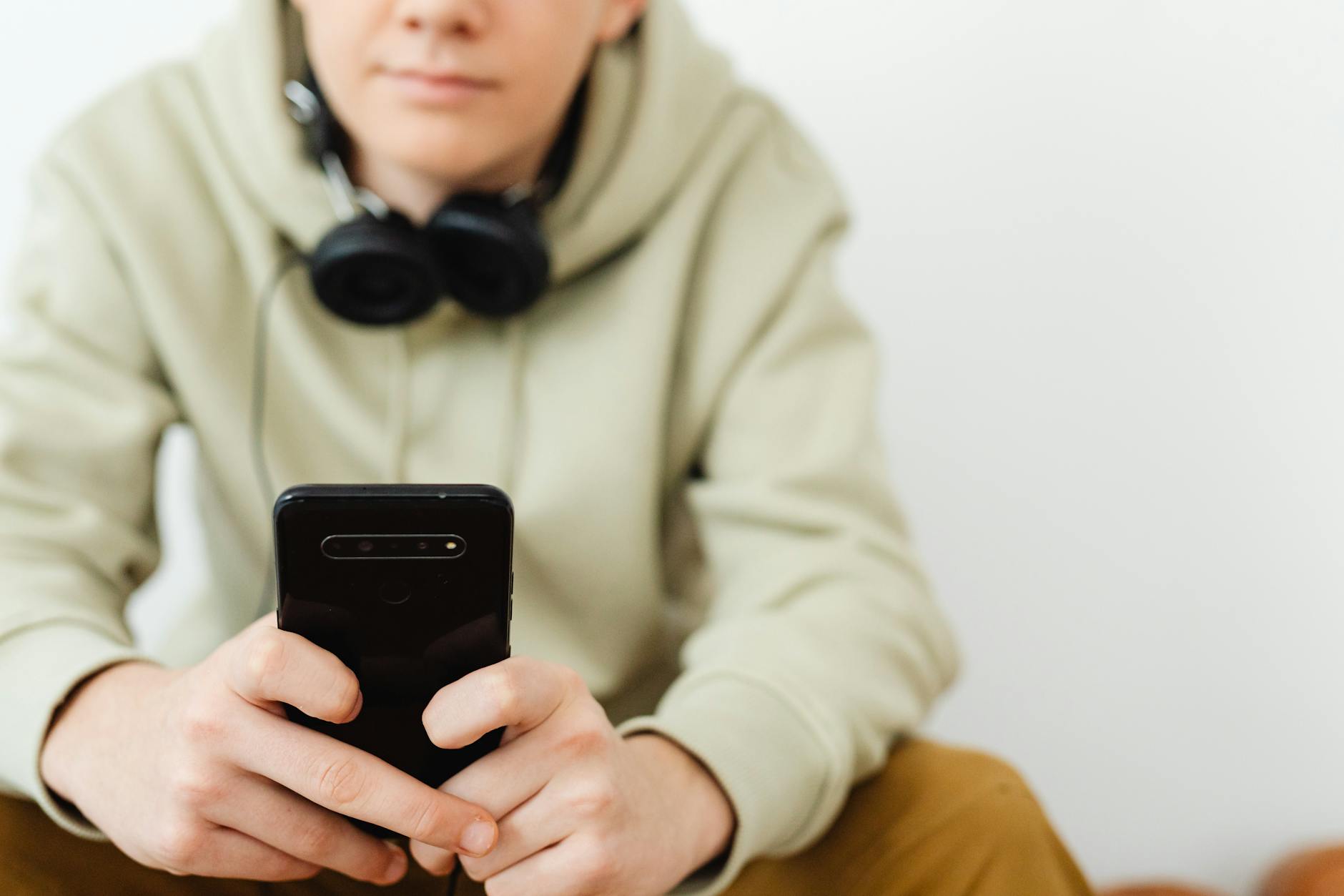 A person wearing a light green hoodie holding a smartphone with headphones around their neck, sitting against a neutral background.