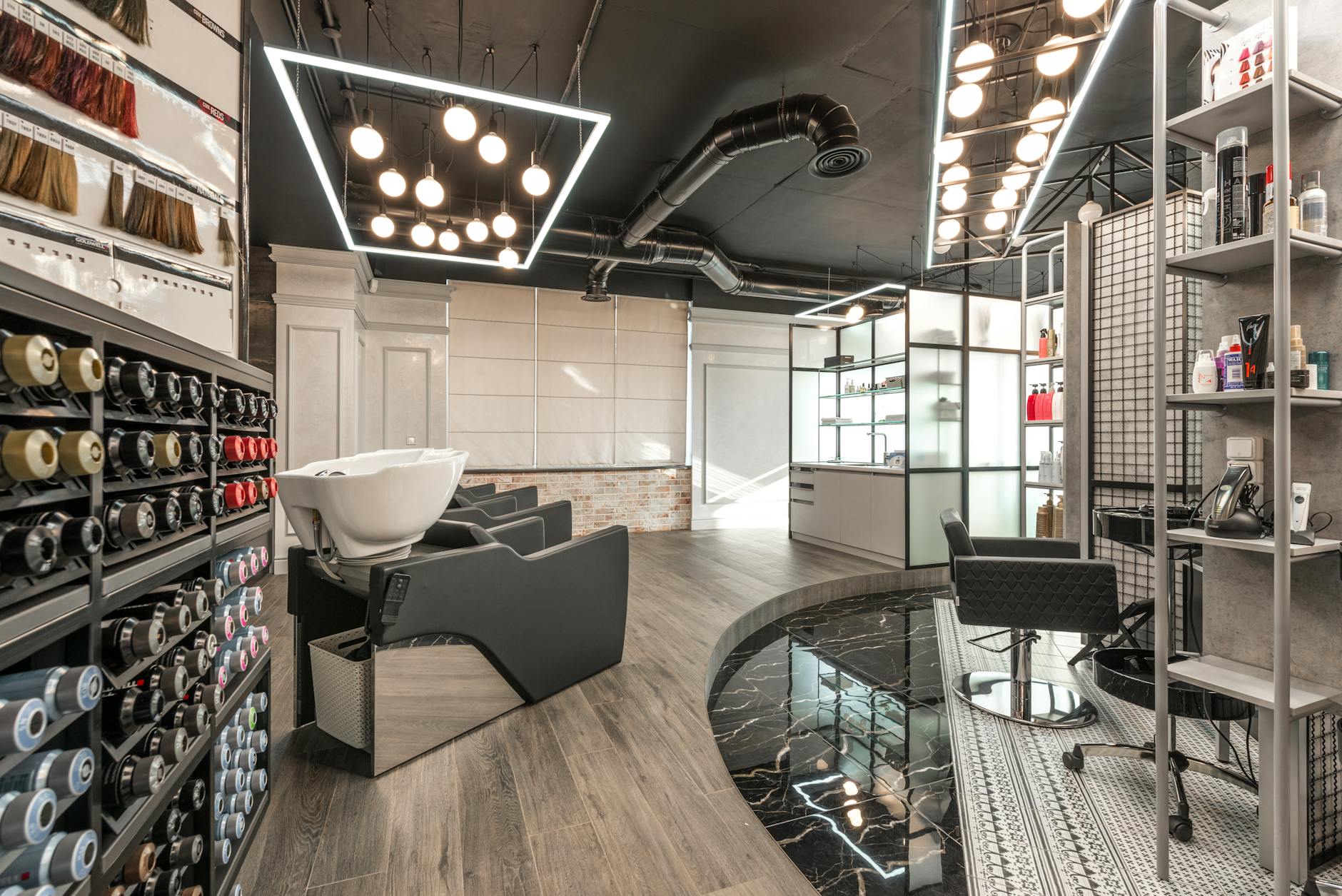 Interior of a modern hair salon featuring a stylish layout with hair treatment stations, a shampoo bowl, and various hair color products displayed on shelves. Ceiling lights illuminate the space, complemented by a sleek design and wooden flooring.
