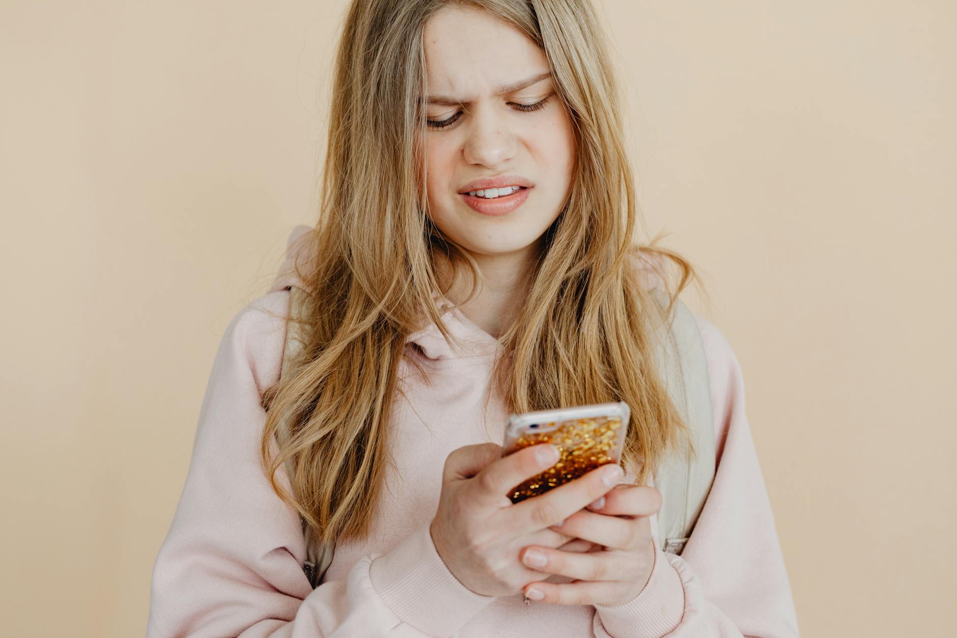 A young woman with long, wavy hair looks at her phone with a confused expression, wearing a light pink hoodie against a beige background.