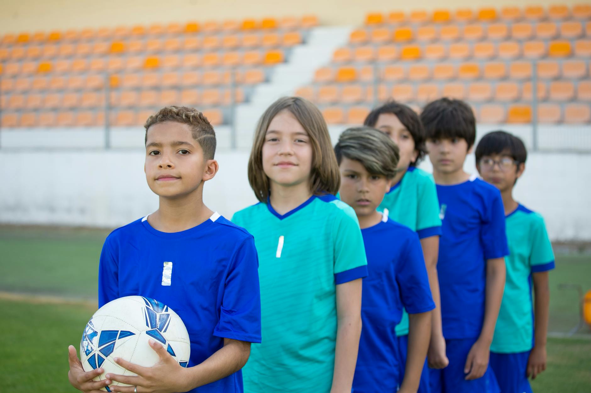 A group of children in soccer uniforms standing in line on a field, with one boy holding a soccer ball in the foreground.