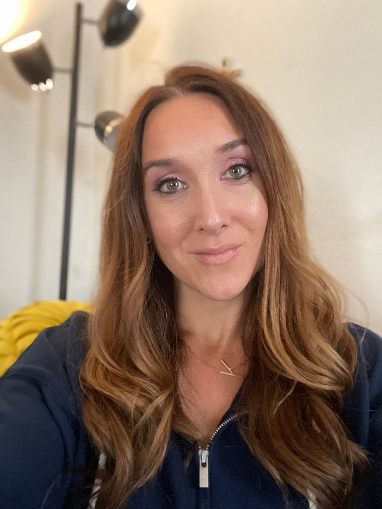 A woman with long, wavy brown hair and natural makeup, smiling at the camera. She is wearing a dark zip-up jacket and a gold necklace, with soft lighting in the background.