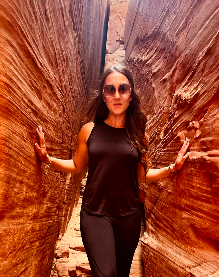 A person wearing sunglasses and a black outfit stands confidently between two red rock formations in a narrow canyon.