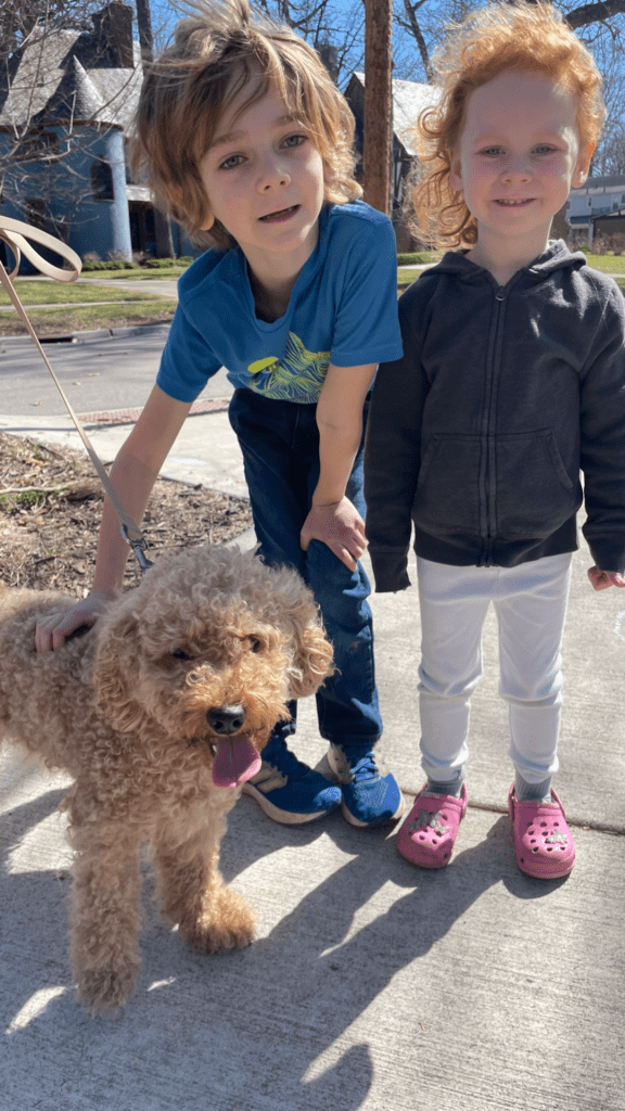 A young boy and girl standing on a sidewalk with a fluffy dog. The boy has light brown hair and is wearing a blue shirt, while the girl has curly red hair and is wearing a dark hoodie. The dog, a tan-colored, curly-haired breed, has its tongue out and is being petted by the boy.