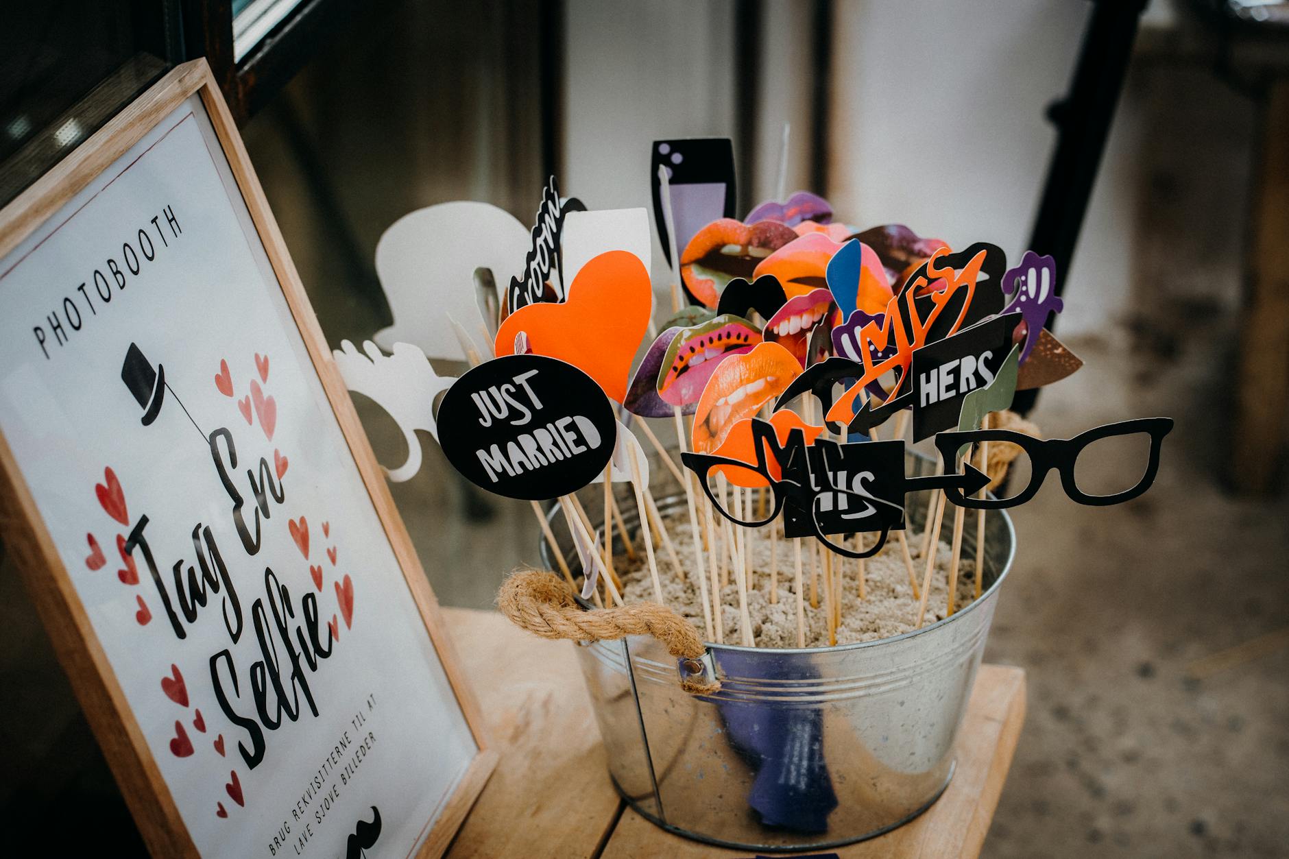 A decorative bucket filled with various photo booth props, including signs that say 'Just Married', 'Mr.', 'Mrs.', and playful shapes like glasses and lips, alongside a framed sign that reads 'Tag En Selfie'.