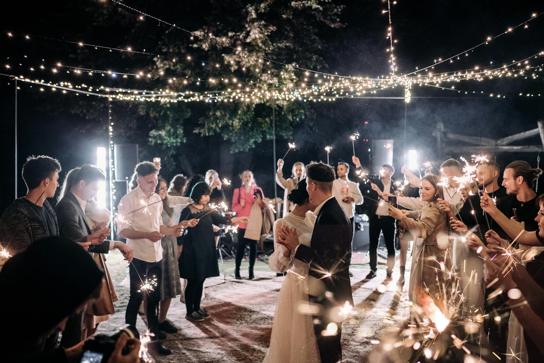 A romantic wedding scene at night with the bride and groom dancing among guests holding sparklers, surrounded by twinkling lights and a festive atmosphere.