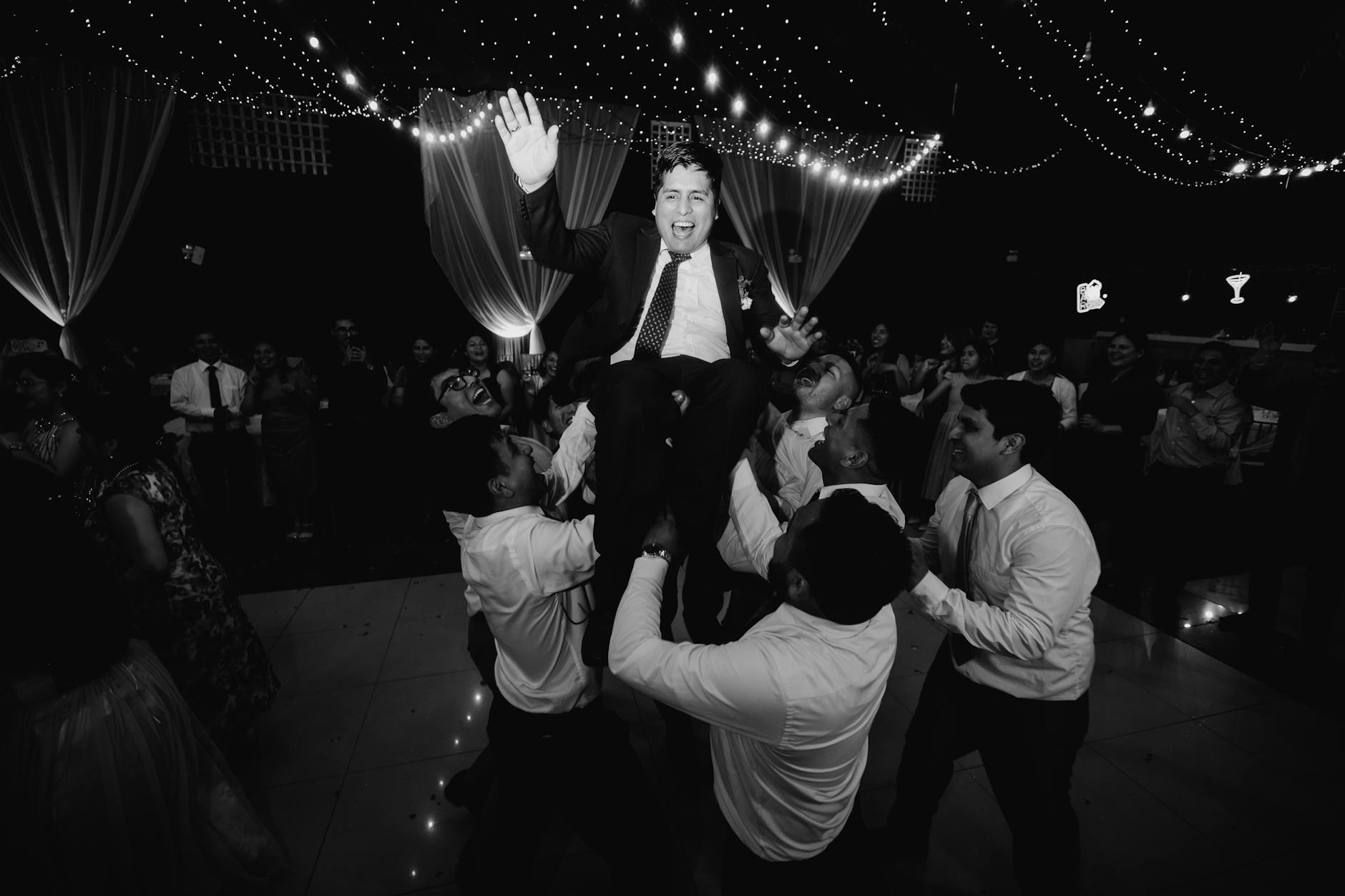 A group of men joyfully lifting a man in a suit while he celebrates, with festive lights in the background.