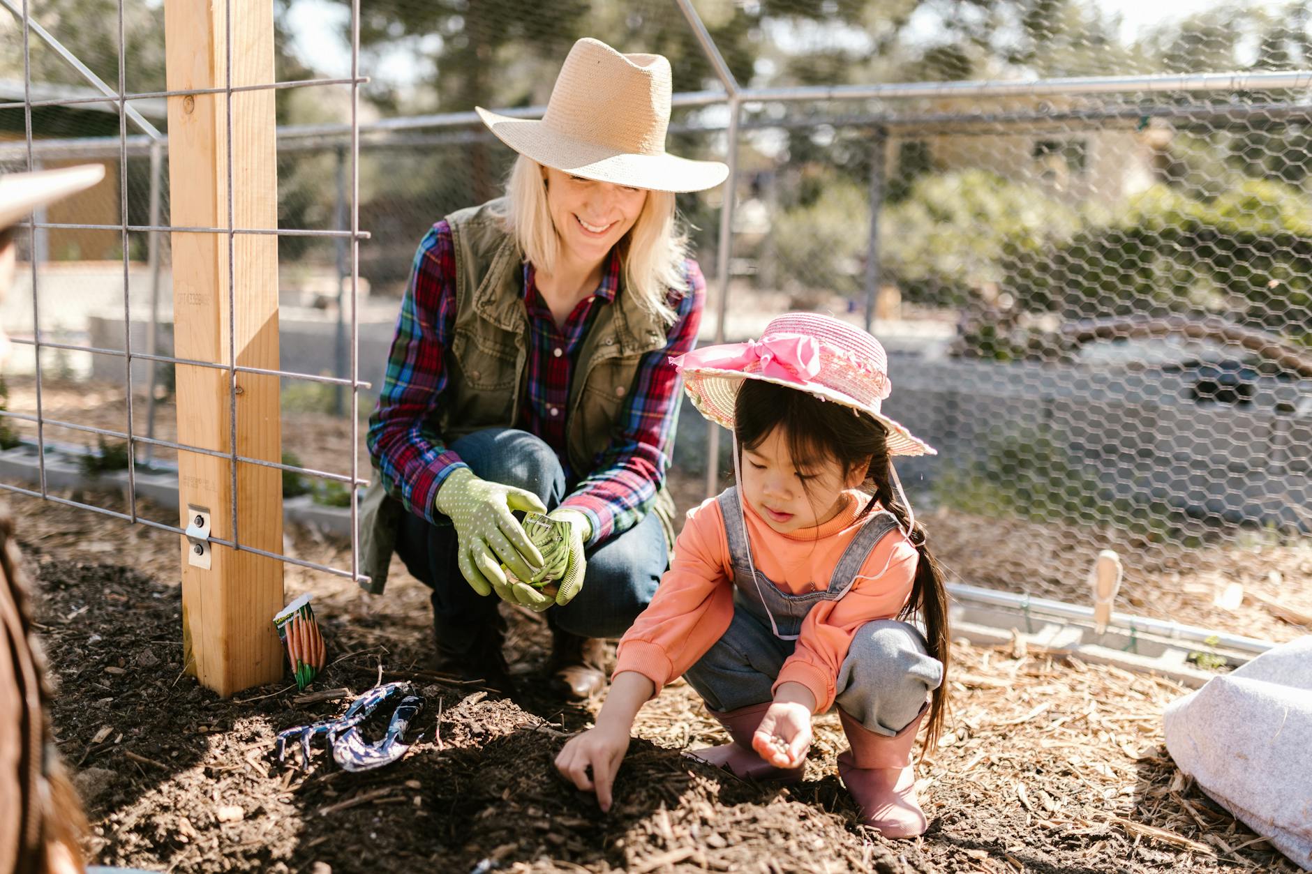 A woman and a young girl gardening together, with the woman kneeling and smiling while the girl digs in the soil, both wearing hats and gloves in a backyard setting.