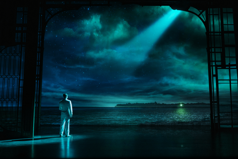 A silhouette of a man in white attire standing on the shore, gazing at a starry night sky and ocean, illuminated by a beam of light.