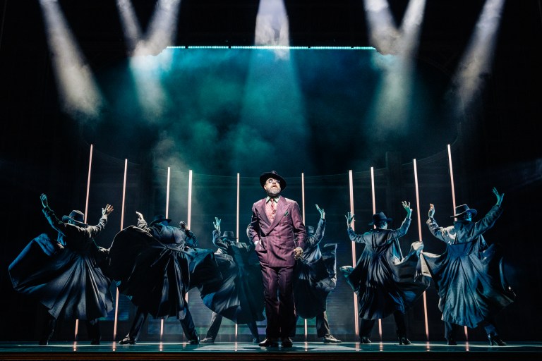 A central performer in a purple suit and hat stands confidently on stage surrounded by dancers in dark cloaks, with dramatic lighting and smoke effects creating an engaging theatrical atmosphere.