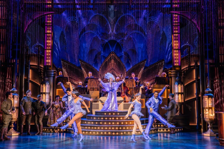 A vibrant stage scene from a musical featuring dancers in blue costumes, with a performer in a grand outfit at the center. The backdrop includes ornate lighting and musicians are visible in the background, creating a lively atmosphere.