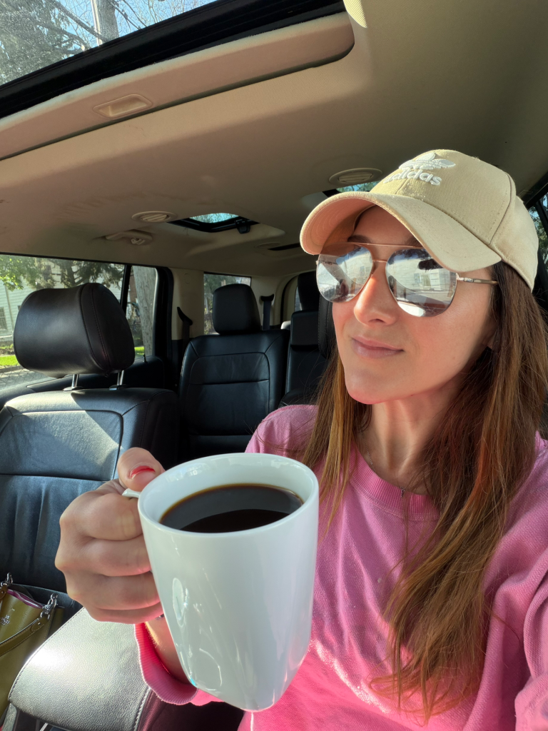 A woman wearing a beige cap and sunglasses sitting in a car, holding a white mug of coffee while wearing a pink sweatshirt.