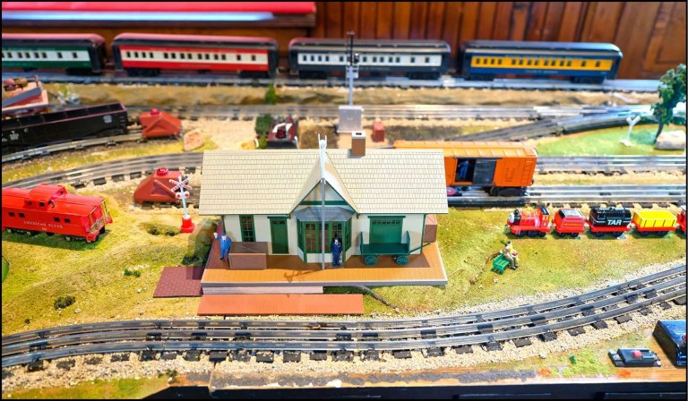 A model train set scene featuring a miniature train station with figures, surrounded by various toy trains and tracks.