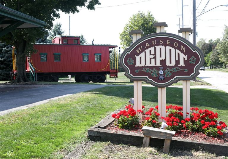 Wauseon Depot sign with a red caboose in the background, surrounded by green grass and flower bed.