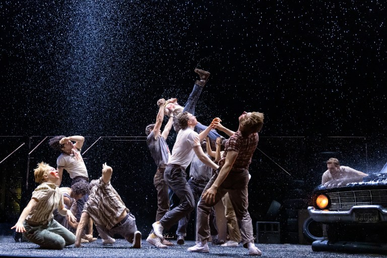 A dynamic theatrical performance scene featuring a group of actors amidst falling water, showcasing various poses and expressions of joy and excitement.