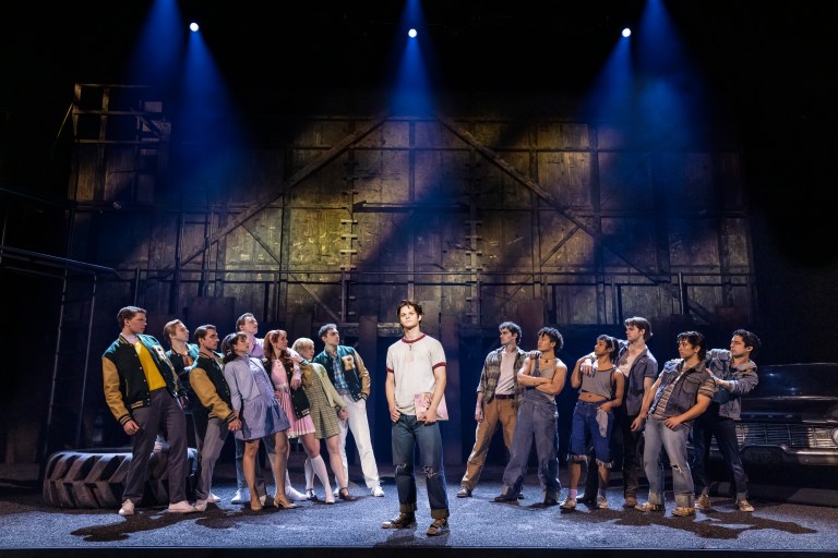 A theatrical performance depicting a group of diverse characters in a dramatic scene, with one character in the foreground standing confidently while others surround him, set against a stylized industrial backdrop with stage lights illuminating the scene.