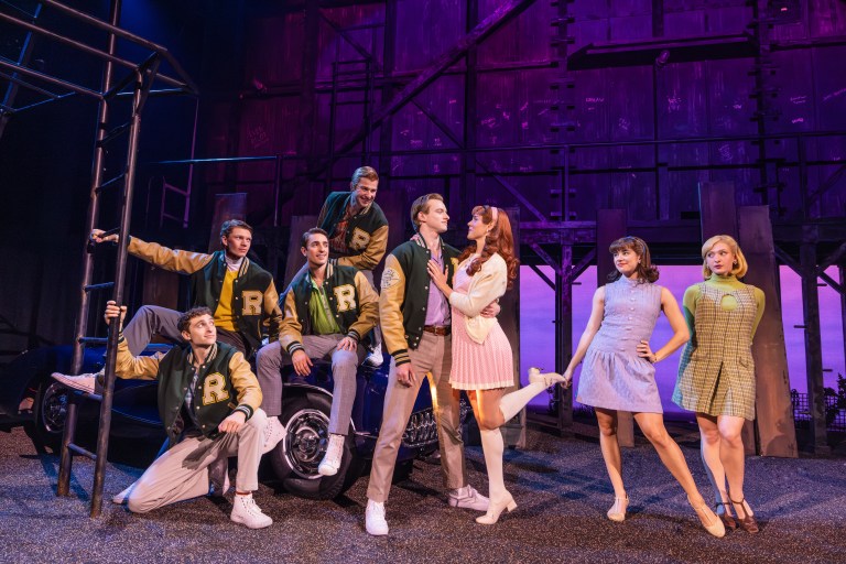 A group of seven performers in a theater production, dressed in colorful costumes. They are posing together on and around a vintage car, with a backdrop illuminated in purple lighting. Some actors are wearing varsity jackets, while others are in casual dresses, creating a dynamic stage presence.