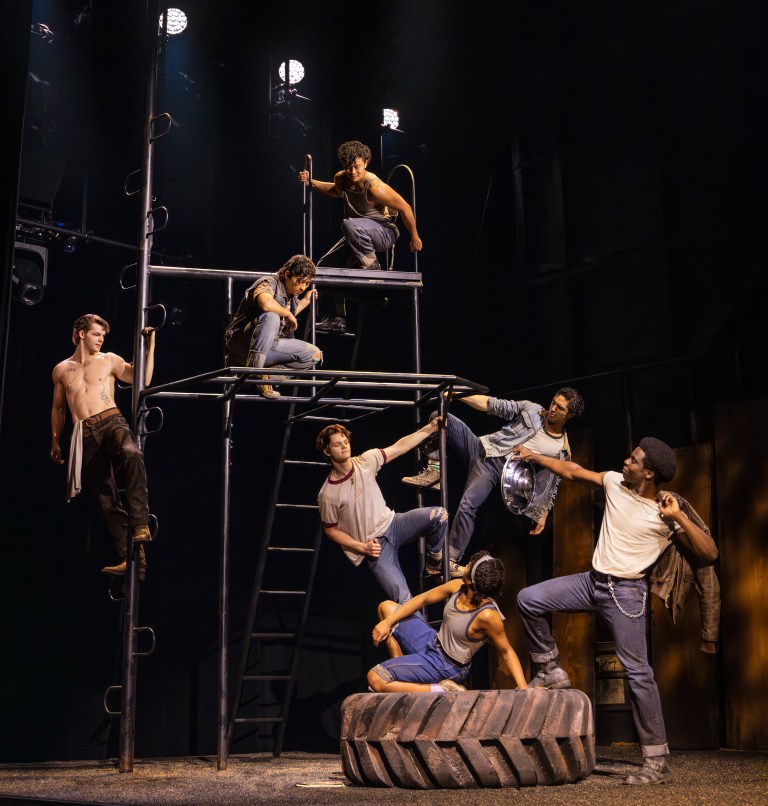 A group of seven performers in a dynamic pose on a set featuring a tall metal structure and a large tire, showcasing a theatrical performance.