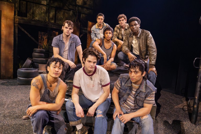 A group of eight young male actors posing together in a theatrical setting, wearing casual and vintage clothing, with a backdrop of worn tires and an industrial environment.
