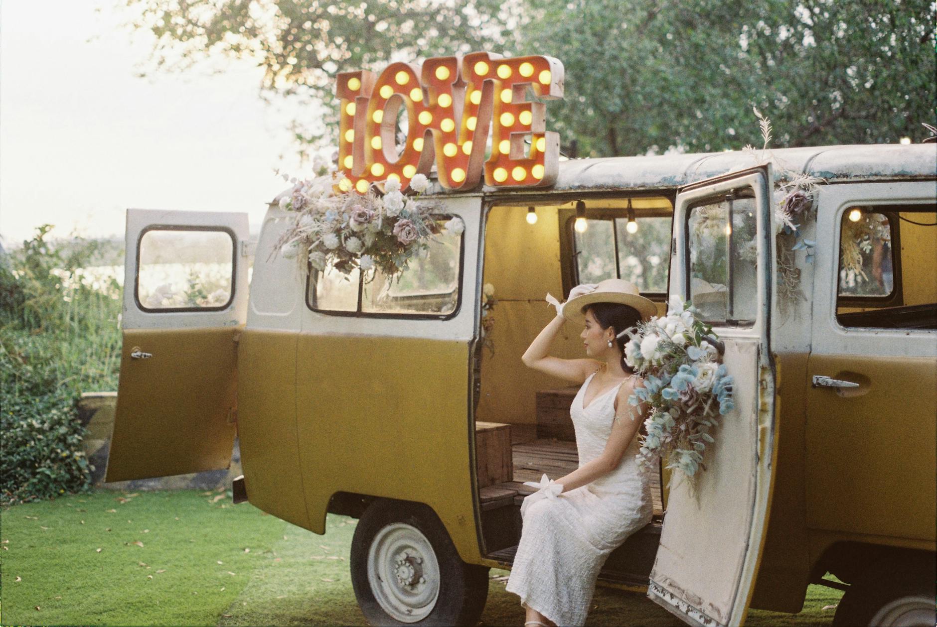 A bride in a white lace dress and wide-brimmed hat sits on the side of a vintage van decorated with flowers and a marquee sign that reads 'LOVE'. The scene is set outdoors with greenery in the background.