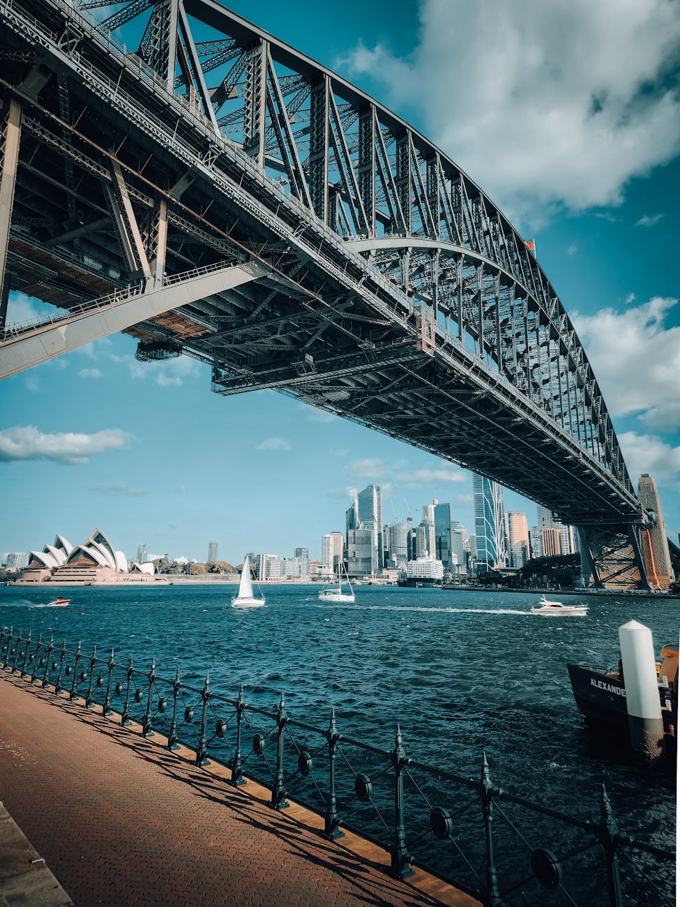 View of the Sydney Harbour Bridge from below, with boats sailing on the water and the Sydney Opera House and city skyline visible in the background.