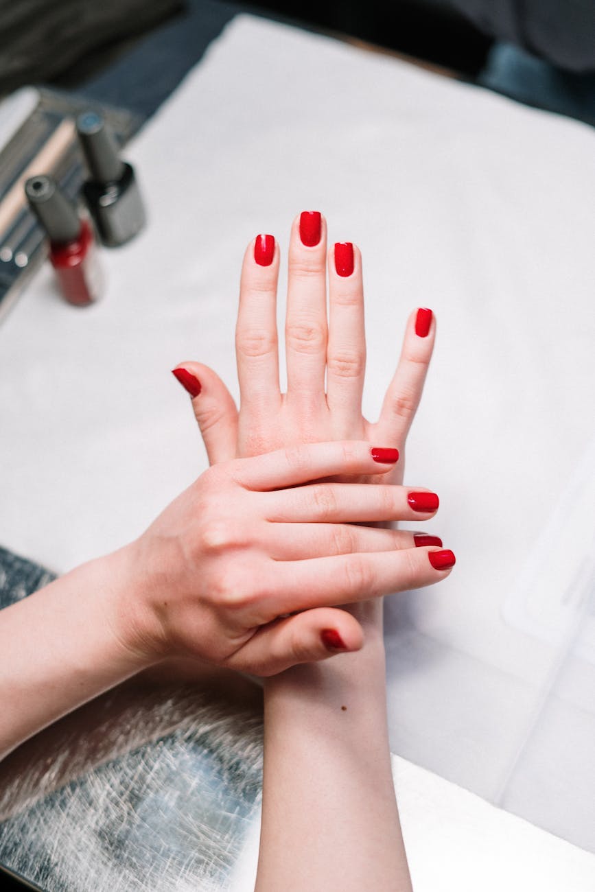 A close-up of two hands with manicured red nails, one hand resting on top of the other, on a white surface with nail polish bottles in the background.