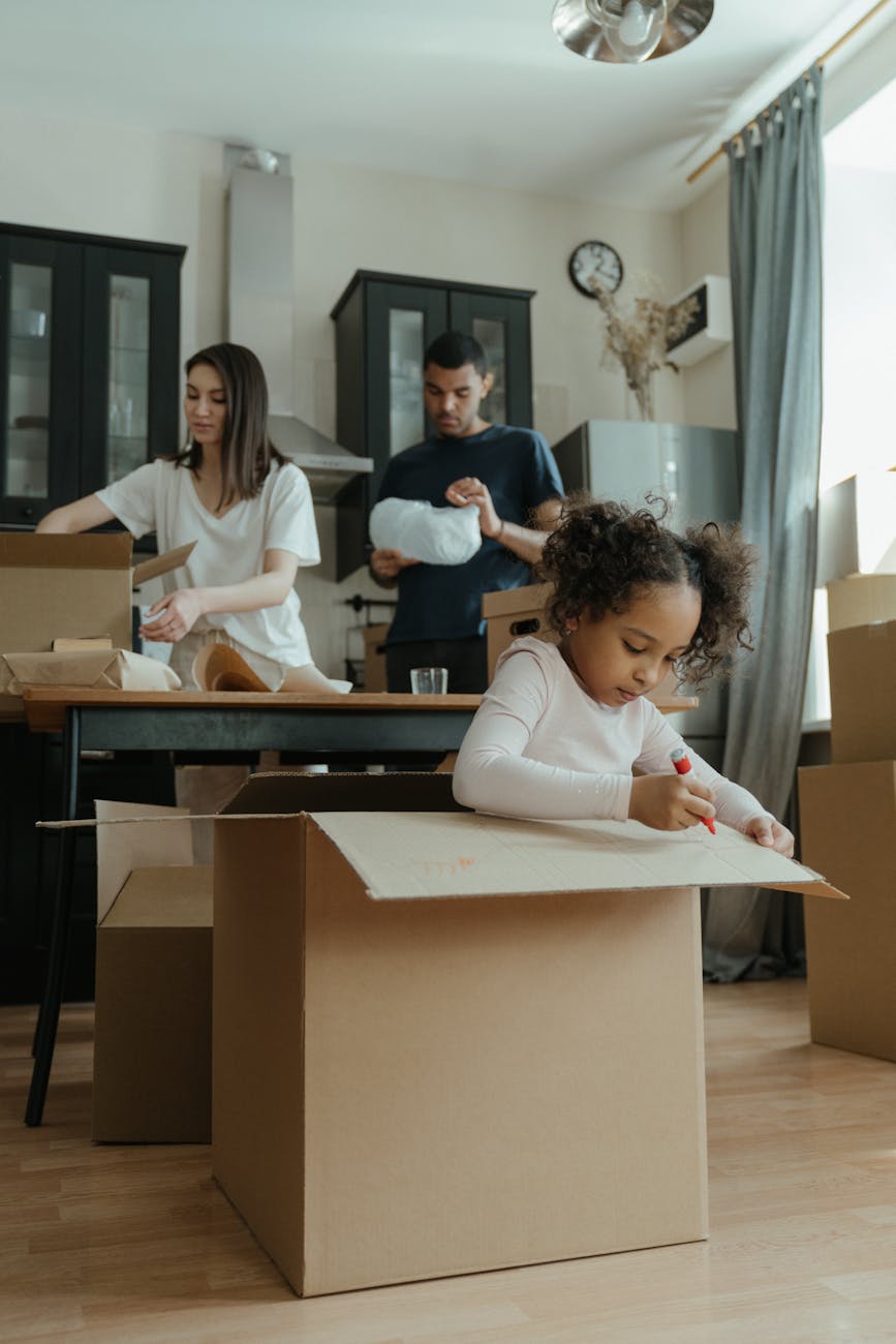 A young girl sitting inside a cardboard box, coloring with a red marker, while two adults are in the background organizing more boxes in a kitchen setting.