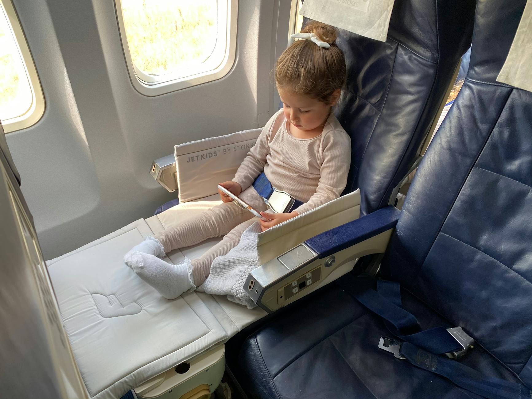 A young child sitting on an airplane seat, using a smartphone while resting their feet on a soft cushion.