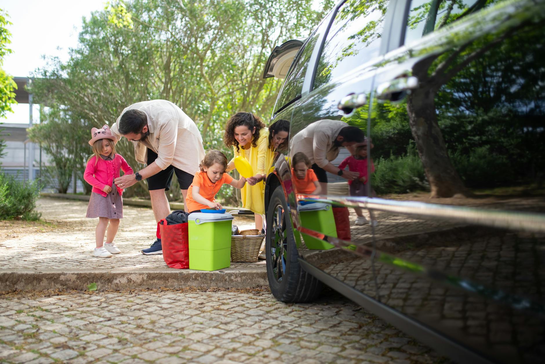 A family unpacking items from a car in a park, with two adults and three children. The adults are interacting with the kids, who are playing and looking at items in colorful containers and bags.