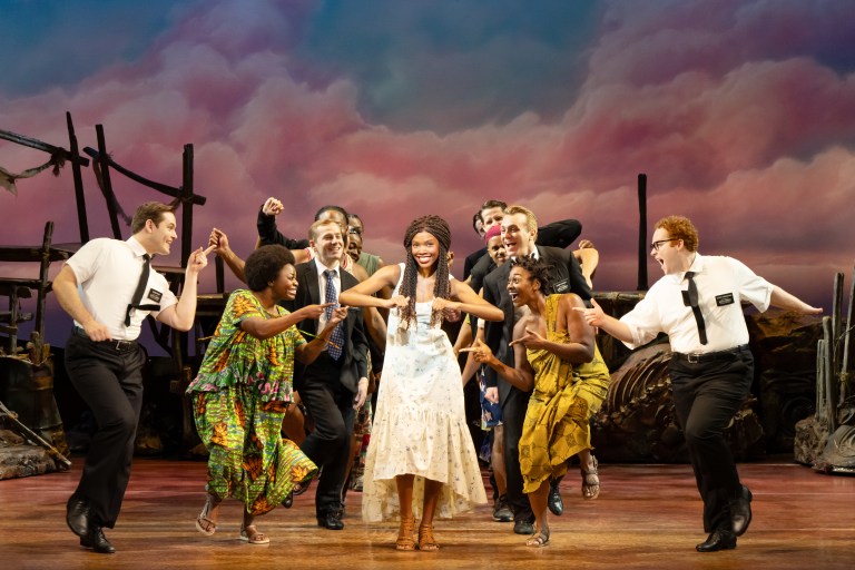 A vibrant musical theater performance featuring a diverse cast, with a central female performer in a white dress surrounded by enthusiastic dancers in colorful outfits, set against a dramatic cloud-filled backdrop.