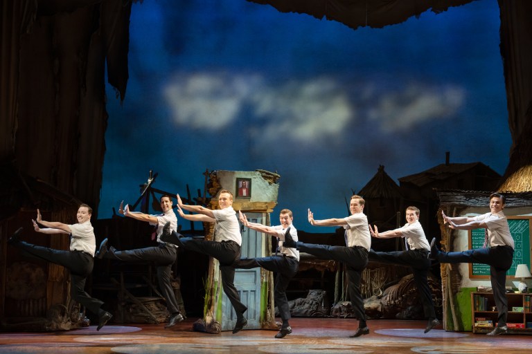 A group of six dancers in white shirts and black pants performing a high-energy dance routine on stage, with a colorful theatrical backdrop.