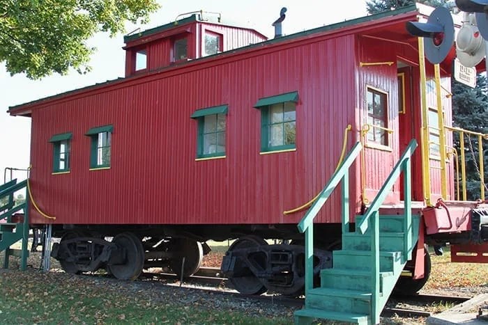 A red train caboose with green trim, featuring multiple windows and a small upper deck, placed on railway tracks with a set of stairs leading to the entrance.