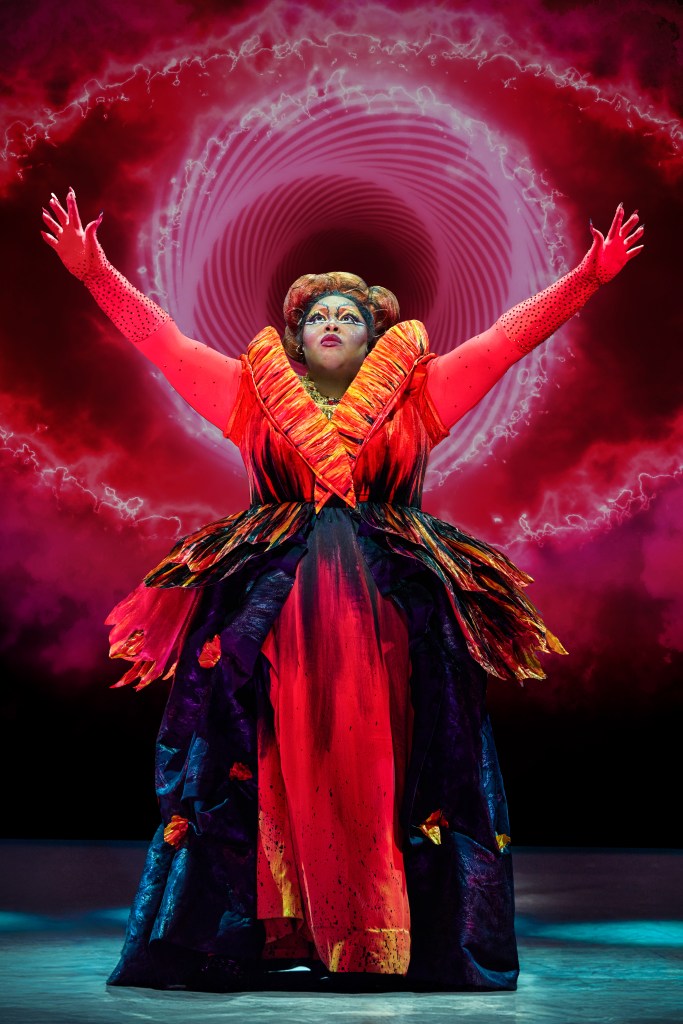 A performer in a vibrant orange and black costume with dramatic sleeves, raising their arms in a powerful pose against a swirling red and purple background.