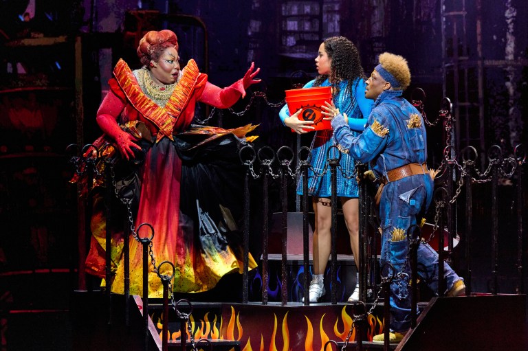 A theatrical scene featuring three performers: a character in a dramatic red costume gesturing expressively, a character in a blue dress holding a container, and another character in blue attire with a light-colored hairstyle, all surrounded by a dark, fiery backdrop.