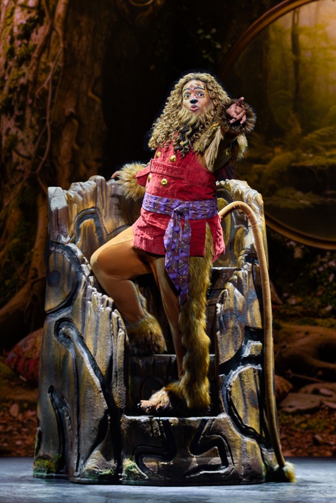A performer dressed as a lion stands on a carved wooden throne, wearing a red outfit with a purple sash and a fluffy mane, gesturing amusingly on stage.