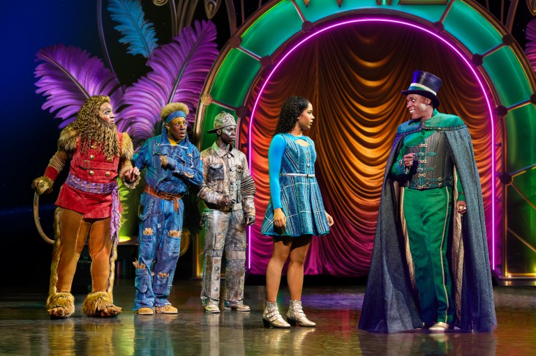 A scene from a theatrical performance featuring five colorful characters in elaborate costumes on stage, with vibrant lighting and a decorative background.