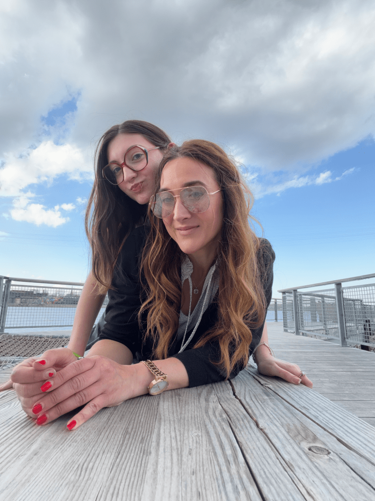 Two women posing on a wooden deck with a scenic view and cloudy sky.