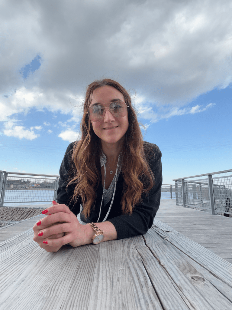 A young woman with long brown hair and sunglasses smiling while leaning on a wooden railing, with a cloudy sky and water in the background.