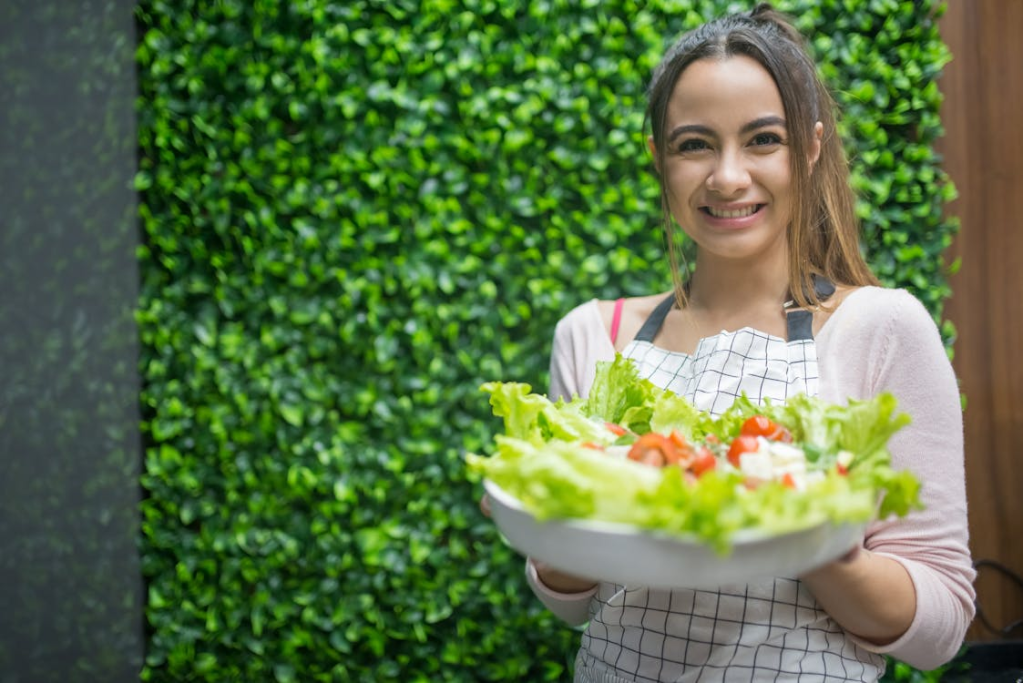 A woman holding a large bowl of fresh salad, smiling in front of a green leafy wall.